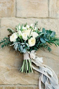 a bouquet of white roses and greenery on a stone wall with ribbon tied around it