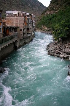 a river running between two buildings next to a mountain side town with green hills in the background