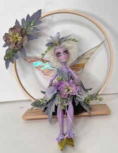 a purple fairy doll sitting in front of a circular frame with flowers and leaves on it