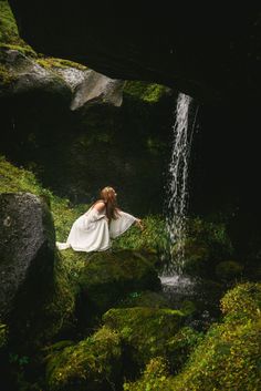 a woman in white dress sitting on mossy rocks next to a waterfall with water coming out of it