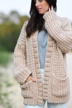 a woman wearing a cardigan sweater and jeans standing in front of the ocean with her hands on her hips