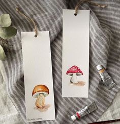 two bookmarks with mushrooms painted on them next to some paintbrushes and a plant