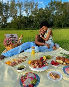 two people sitting on a blanket in the grass with plates of food and drinks around them