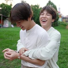 two young men standing next to each other in the grass smiling at something on his cell phone