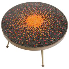 a round mosaic table with metal legs and an orange center piece on the top, sitting in front of a white background