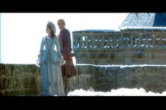 two people standing next to each other in front of a stone wall with snow on it