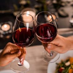 two people toasting with red wine in front of a plate of salad and candles