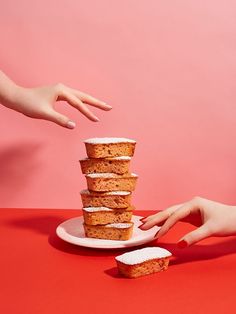 two hands reaching for a stack of cake on a white plate against a pink background