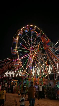 people walking around an amusement park at night with lights on the ferris wheel in the background