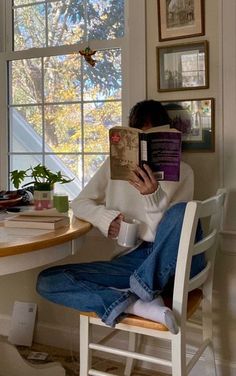 a woman sitting at a table reading a book and drinking coffee in front of a window