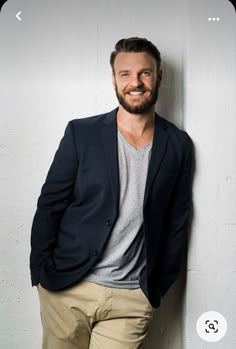 a man leaning against a white wall with his hands in his pockets and smiling at the camera