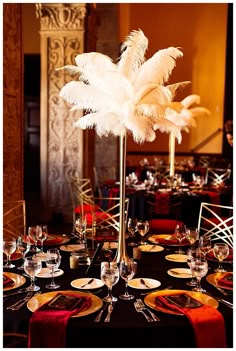 a tall centerpiece with white feathers sits on top of a table set for an event