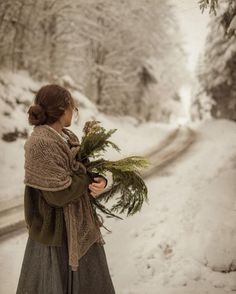 a woman standing in the middle of a snow covered road holding a bunch of plants