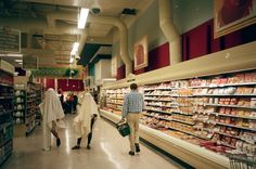 two people are walking down the aisle of a grocery store while others shop in the aisles