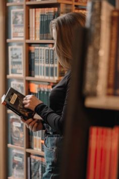 a woman standing in front of a bookshelf holding a book and looking at it