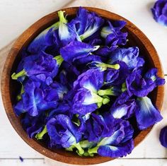 a wooden bowl filled with purple flowers on top of a white table next to blue petals