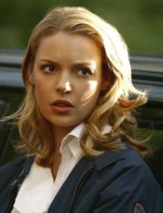 a woman with blonde hair wearing a blue jacket and white shirt is standing in front of a car