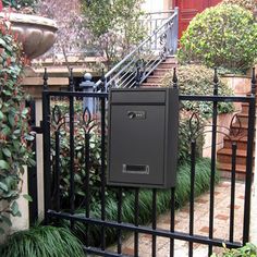 a mailbox on the side of a black iron fence in front of some bushes