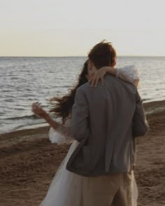 a bride and groom hug on the beach as the sun sets in the distance behind them