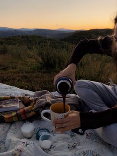 a woman sitting on top of a blanket pouring something into a cup with a spoon