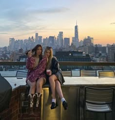 two young women sitting on top of a building in front of a city skyline at sunset