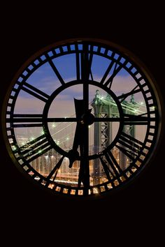 a large clock on the side of a building with a view of a bridge in the background