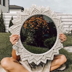 a woman sitting on the grass holding up a mirror with flowers in front of her