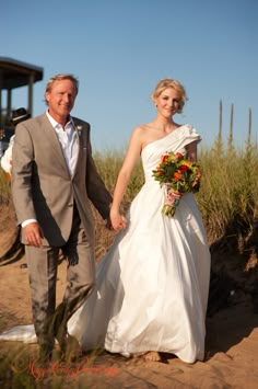 a bride and groom holding hands walking on the beach with their bouquets in hand