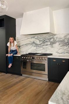 a woman sitting on the counter in a kitchen with black cabinets and marble backsplash