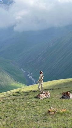 a man standing on top of a lush green hillside next to a valley filled with mountains