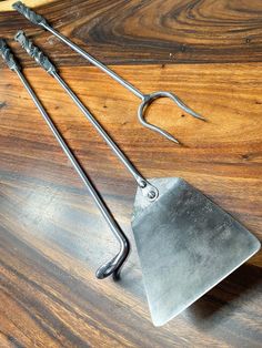 three metal tools are laying on a wooden table top, one has a hook and the other is a pair of tongs