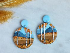 a pair of earrings with trees and mountains painted on them, sitting next to dried grass