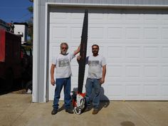 two men standing next to each other in front of a garage holding up a large black object