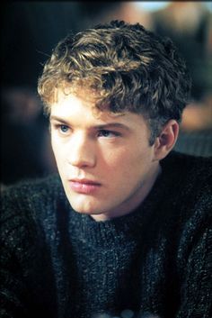 a young man with curly hair wearing a black sweater and looking at the camera while sitting down