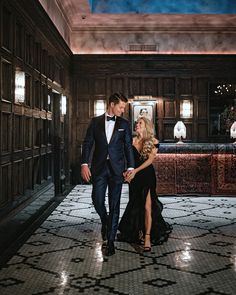 a man and woman in formal wear walking through an ornate hallway with lights on the ceiling