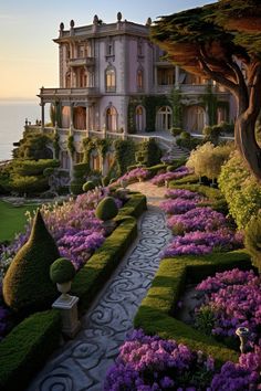 a large house with lots of flowers and bushes in front of the water at sunset