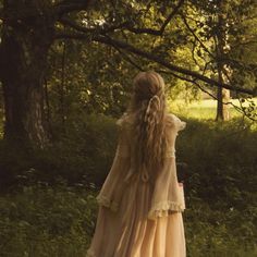 a woman with long hair in a dress walking through the grass near trees and bushes