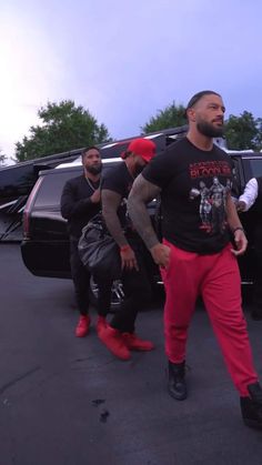 two men in red pants and black t - shirts are walking towards a car