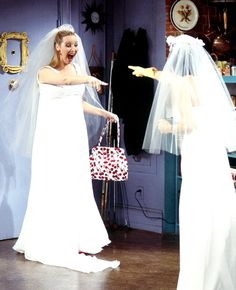 two women dressed in wedding attire, one pointing at the bride's handbag