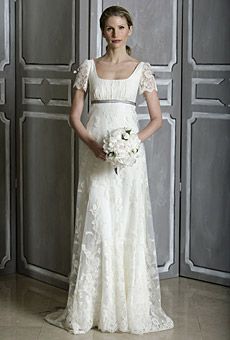 a woman standing in front of a wall wearing a white dress and holding a bouquet