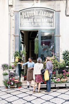 three women are standing in front of the entrance to a flower shop with potted plants