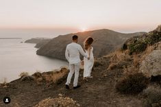 a man and woman holding hands while standing on top of a hill next to the ocean