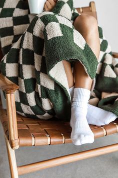 a woman sitting in a chair with a cup of coffee on her lap, wrapped in a green and white checkered blanket