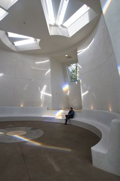 a person sitting on a bench in a room with white walls and ceilings that have sunlight streaming through the windows