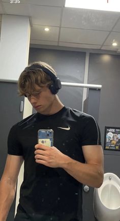 a young man wearing headphones looking at his cell phone while standing in front of a urinal