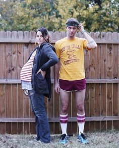 a man and woman standing next to each other in front of a fence with trees