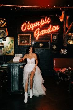 a woman in a white dress sitting at a table with a cake on it and an orange neon sign behind her