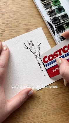 two hands are holding a business card on top of a wooden table next to some paintbrushes