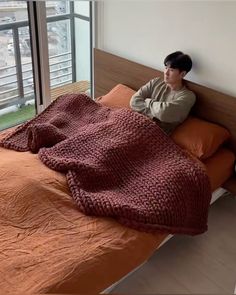 a man sitting on top of a bed next to a window with an orange blanket