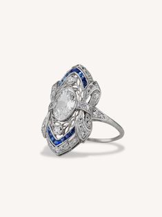 This antique Edwardian ring in motif and craftsmanship is iconically early 20th century. Centered with a GCAL certified .85 carat moval shaped diamond with H-I color and VS2 clarity. Enhanced by 44 full and single cut diamonds. Calibré cut sapphire accents add contrast and dimension. Crafted in platinum with charming bow details on the shoulders and a delicate openwork design adorned with foliate filigree. Everything about this ring exudes ultra-femininity and delicacy making it on trend for mot Luxury Blue Elegant Filigree Ring, Luxury Antique Rings With Intricate Design, Luxury Blue Filigree Ring For Engagement, Luxury Victorian White Gold Sapphire Ring, Luxury Blue Classic Filigree Ring, Luxury Antique Rings For Evening, Edwardian Engagement Ring Erstwhile Jewelry, Antique Rings Edwardian, Rings Edwardian